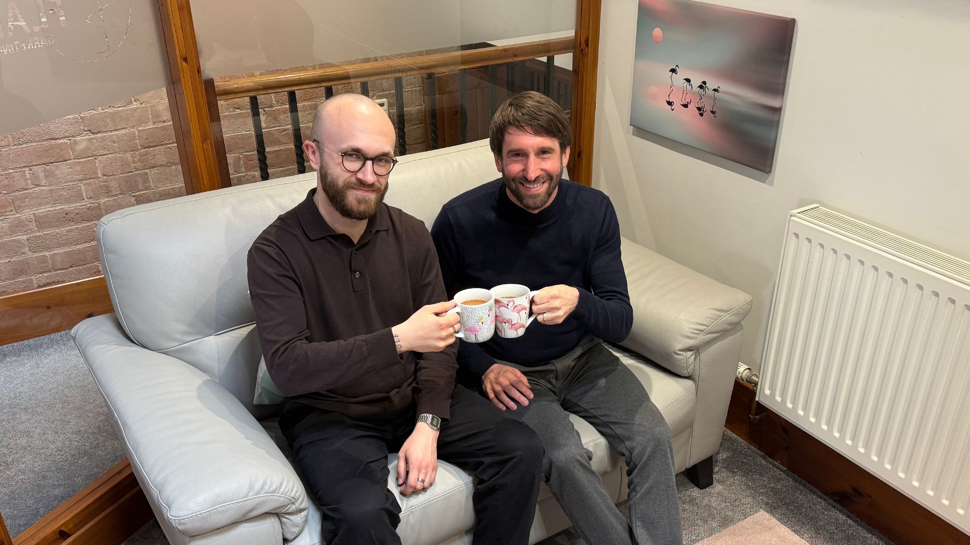 George and Rich holding flamingo-themed mugs, sitting on a grey sofa in the office.